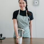 Positive young Asian woman wearing apron giving paper cup with coffee to go while standing at wooden counter during work in modern coffee house and looking at camera with smile