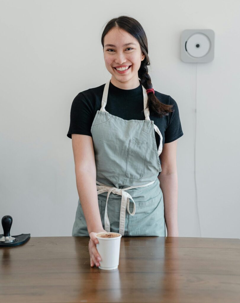 Positive young Asian woman wearing apron giving paper cup with coffee to go while standing at wooden counter during work in modern coffee house and looking at camera with smile