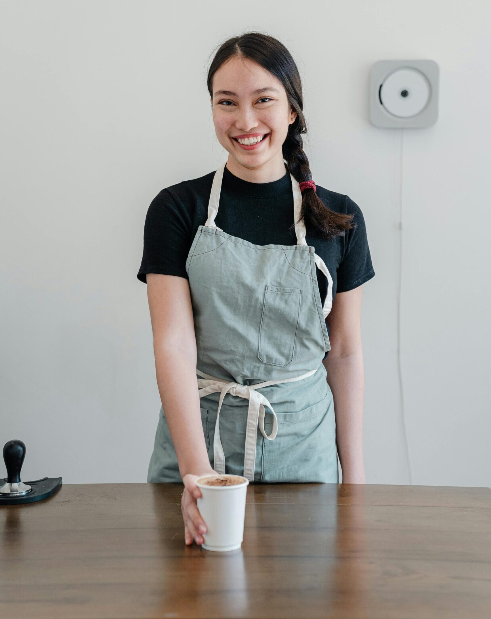 Positive young Asian woman wearing apron giving paper cup with coffee to go while standing at wooden counter during work in modern coffee house and looking at camera with smile