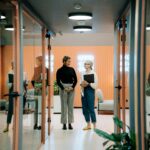Two women discuss business in a stylish office hallway with glass doors and modern decor.