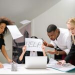 Team of stressed colleagues in an office dealing with paperwork chaos, asking for help.